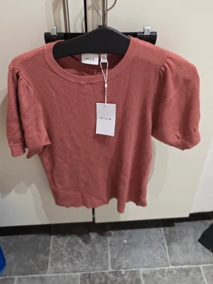 Rostfärgad ribbad topp från Vila - Trendig rostfärgad ribbad topp från Vila med puffiga korta ärmar och rund halsringning. Materialet är mjukt och stretchigt, perfekt för dig som gillar en bekväm men snygg stil. Enkel att matcha med både jeans och kjol.
