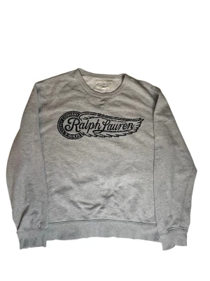 Grå sweatshirt från Ralph Lauren - Grå sweatshirt från Ralph Lauren Denim & Supply med svart tryck framtill. Tröjan har rund halsringning, ribbade muddar och avslappnad passform. Perfekt för en casual stil och enkel att matcha med jeans eller joggers. Den är i storlek xl i ungdom så passar m