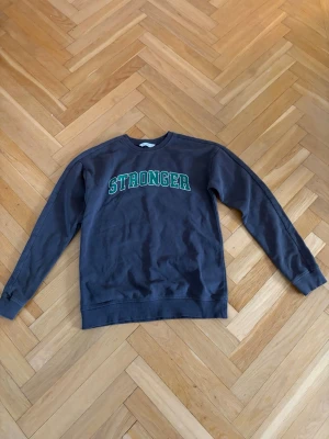 Mörkgrå sweatshirt med STRONGER-tryck - Mörkgrå sweatshirt från STRONGER med grönt och vitt tryck på bröstet. Klassisk rund halsringning och långa ärmar. Tillverkad i mjukt bomullsmaterial för en avslappnad och bekväm stil.