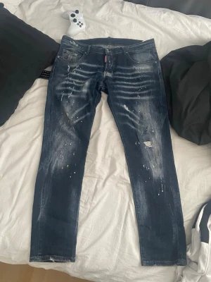 Mörkblå distressed jeans med färgstänk - Säljer ett par mörkblå jeans med slitningar, färgstänk och en patchad detalj på ena benet. Jeansen har en rak passform och klassiska fem fickor. Perfekta för dig som gillar en edgy och avslappnad stil.