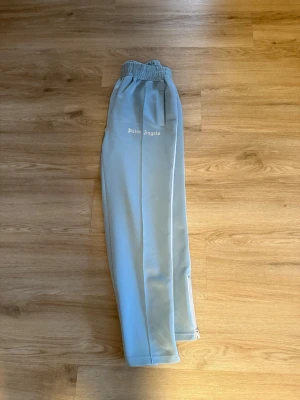 Palm Angels ljusblå mjukisbyxor - Snygga ljusblå mjukisbyxor från Palm Angels med vita sidoränder och elastisk midja. Byxorna har raka ben, fickor och logga tryckt på ena sidan. Perfekta för en chill och trendig look. Perfekt för dig som är runt 180 