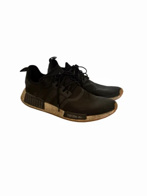 Svarta Adidas NMD - Svarta Adidas NMD_R1 sneakers med klassiska tre ränder på sidan, stickad ovandel i mesh och bruna Boost-sulor. Skorna har svarta snören och detaljerade hälflikar med Adidas-logga och text. Perfekta för en streetwear-look.