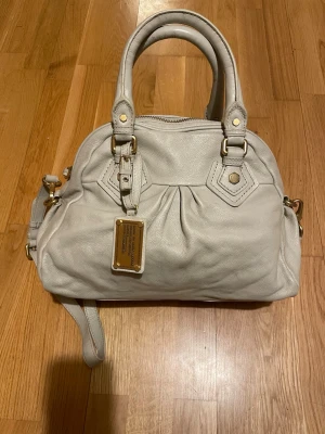 Ljusgrå Marc by Marc Jacobs handväska - Snygg ljusgrå handväska från Marc by Marc Jacobs i mjukt läder. Väskan har dubbla handtag, avtagbar axelrem och guldfärgade metalldetaljer. Framtill sitter en metallbricka med logotyp. Perfekt för dig som gillar stilrena accessoarer.