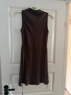 Brun ärmlös klänning i satin - Snygg brun ärmlös klänning i satin med lätt glansig finish och markerad midja. Klänningen har en enkel, elegant design och faller mjukt längs kroppen. Från BikBok, använd 1 gång