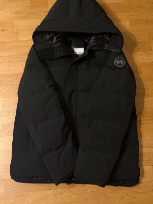 Svart dunjacka från Canada Goose - Snygg svart dunjacka från Canada Goose med huva och klassisk logotyp på ärmen. Jackan har en stilren design, dragkedja och är perfekt för kalla vinterdagar. Materialet är slitstarkt och håller värmen riktigt bra. Helt ny