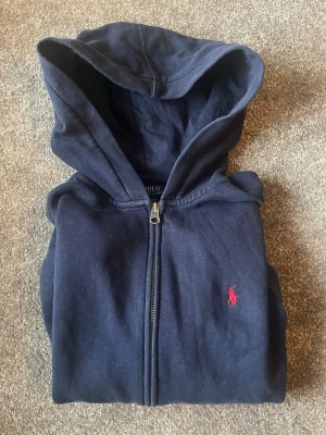 Mörkblå hoodie från Polo Ralph Lauren, st. 10-12 - Mörkblå hoodie med dragkedja från Polo Ralph Lauren, storlek 10-12. Klassisk design med huva och röd broderad logga på bröstet.