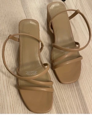 Beige sandaler med blockklack från H&M - Stilrena beige sandaler från H&M med tunna remmar och fyrkantig tå. Sandalerna har en låg blockklack och är tillverkade i syntetmaterial. Perfekta för en trendig och minimalistisk look.