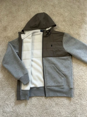 Grå hoodie från Polo Ralph Lauren - Säljer en grå hoodie från Polo Ralph Lauren med kontrasterande mörkgrått parti över bröstet och huva. Dragkedja framtill, två fickor och mjukt vitt fleecefoder på insidan. Klassisk logga broderad på bröstet. Perfekt för chill dagar eller när du vill vara extra bekväm.