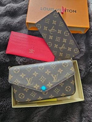 Monogram handväska - Säljer en klassisk handväska i brunt monogrammönster med guldiga detaljer och röd insida. Väskan har en avtagbar kedjerem, flera fack och kommer med matchande plånbok och korthållare. Dustbag + box mefföljer.