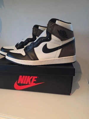 Nike Air Jordan 1 High retro mocha - Nike Air Jordan 1 High mocha sneakers i svart, vitt och brunt med klassisk Swoosh-logga på sidan. Ovandel i läder och mocka, högt skaft och ikonisk Air Jordan Wings-logga vid ankeln. Snygg och tidlös modell som passar till streetwear. Nya i orginalkartong och även kartongen som skickades från Nike.