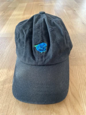 Svart keps med blå rosbrodyr - Svart keps i klassisk modell med en broderad blå ros framtill. Kepsen har böjd skärm och justerbar passform baktill. Tillverkad i bomullstyg och passar perfekt för en avslappnad streetstil.