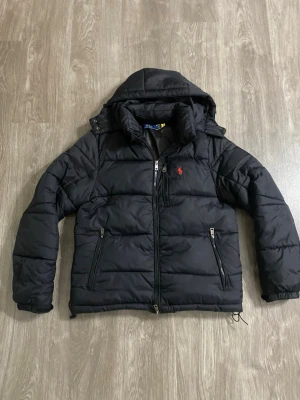 Svart pufferjacka från Polo Ralph Lauren - Snygg svart pufferjacka från Polo Ralph Lauren med röd logga på bröstet. Jackan har huva, två dragkedjeförsedda fickor och dubbla dragkedjor framtill. Perfekt för kalla dagar och riktigt skön att bära. Klassisk design som funkar till det mesta.