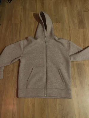 Beige zip hoodie med huva - Säljer en stilren beige hoodie med dragkedja framtill och två stora fickor. Hoodien har en klassisk huva och är gjord i ett mjukt, tjockt material som känns riktigt skönt. Perfekt för dig som gillar en enkel och clean look.