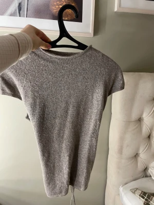Grå stickad topp med knyt i ryggen - Säljer en grå/brun, kortärmad stickad topp med rund hals och snygg knytning i ryggen. Perfekt till jeans eller kjol för en chill och trendig look. Materialet är mjukt och stretchigt för extra komfort. Använd en gång och tvättad efter det☁️