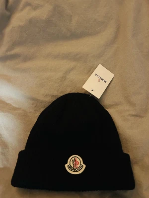 Svart ribbstickad mössa Moncler - Snygg svart ribbstickad mössa från Moncler med uppvikt kant och klassisk Moncler-logga framtill. Tillverkad i mjukt material som håller värmen och ger en clean look. Perfekt accessoar för kalla dagar och enkel att matcha med olika outfits.