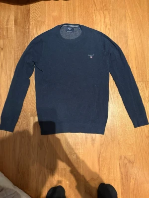 Mörkblå tröja från GANT - Klassisk mörkblå rundhalsad tröja från GANT med diskret logga broderad på bröstet. Tröjan har långa ärmar och ribbade muddar vid ärmslut och nederkant. Tillverkad i mjukt och tunt material som passar perfekt till lager på lager. Är en d men är en liten s 