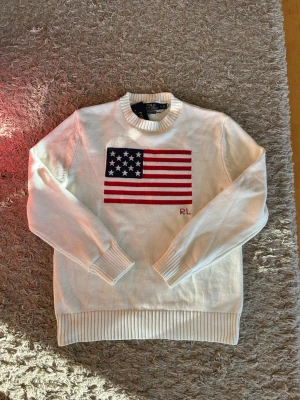 Vit stickad tröja från Polo Ralph Lauren - Vit stickad tröja från Polo Ralph Lauren med amerikansk flagga i rött, vitt och blått på bröstet samt RL-broderi. Tröjan har rund halsringning, ribbade muddar och nederkant. Perfekt för dig som gillar klassisk och preppy stil.
