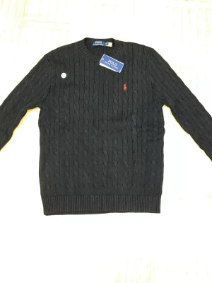 Svart kabelstickad tröja Polo Ralph Lauren - Kabelstickad svart tröja från Polo Ralph Lauren med klassisk röd logga broderad på bröstet. Tröjan har rund halsringning, ribbade muddar och är tillverkad i mjukt material. Perfekt för dig som gillar stilrena och tidlösa plagg.