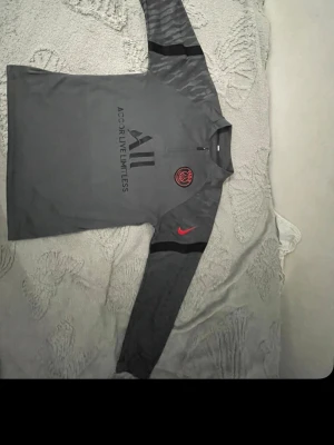 PSG långärmad träningströja Nike - Mörkgrå långärmad träningströja från PSG med svarta och röda detaljer. Tröjan har halv dragkedja, PSG-logga och Nike Swoosh på bröstet samt texten 'Accor Live Limitless' framtill. Materialet är troligtvis polyester, perfekt för träning.