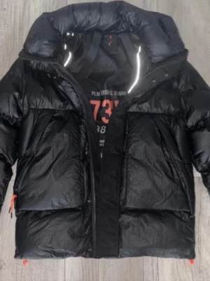 Svart pufferjacka med huva - Snygg svart pufferjacka från PLM RIGID DENIM med stor huva och två rejäla framfickor. Jackan har glansigt material och dragkedja framtill. Perfekt för kalla dagar när du vill hålla värmen och ändå se cool ut.