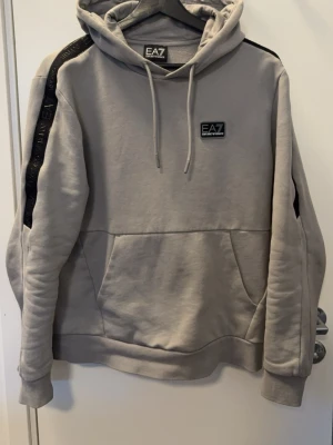 Grå hoodie från EA7 Emporio Armani - Snygg grå hoodie från EA7 Emporio Armani med svarta detaljer på axlarna och logga på bröstet. Hoodien har dragsko i huvan och en stor magficka. Perfekt för en avslappnad och trendig stil.