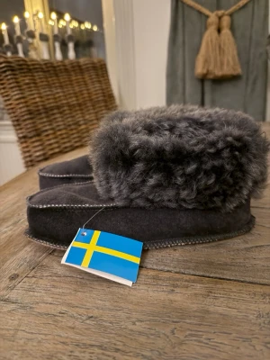 Mörkgrå fårskinnstofflor Lammhagen - Mysiga mörkgrå tofflor från Lammhagen i äkta fårskinn. De har en fluffig, mjuk kant och handsydda detaljer längs sulan. Perfekta för att hålla fötterna varma hemma. Svensk design med naturligt material och klassisk look. Ha åtminstone en centimeter tillgodo så att sömmarna ej nöts. Handsydda i Sverige. Oanvända.