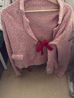 Odd Molly Rosa mönstrad kofta med rosett - Odd Molly en rosa och vit mönstrad kofta med dekorativ rosett framtill. Koftan har lång ärm, liten ficka på ena sidan och en avslappnad krage. Perfekt för dig som gillar unika detaljer och sköna material.