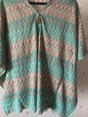 Pernilla Wahlgrens stickad poncho i turkos och beige - Snygg stickad poncho med kort ärm och dragkedja framtill. Mönstret är i turkos, beige och inslag av rosa, med glansig finish. Perfekt att slänga över en topp för en cool look.