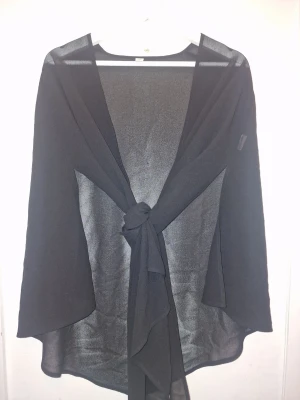 Svart transparent knytkofta - Snygg svart transparent bolero med knytband framtill. Långa ärmar och lätt, luftig känsla som passar perfekt över ett linne eller topp. Materialet är tunt och ger en elegant look. Passar S-M. Inköpt i New York.