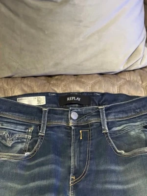 Replay Anbass blå jeans - Snygga blå jeans från Replay, modell Anbass. Jeansen har en klassisk femficksdesign, med slitningar och ljusa tvättade partier för en cool look. De är raka i modellen och har detaljerade sömmar. Perfekta för en avslappnad streetstil.