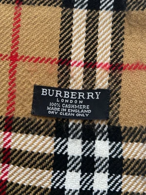 Burberry beige rutig cashmerehalsduk - Klassisk beige halsduk från Burberry i 100% cashmere med det ikoniska rutmönstret i svart, vitt och rött. Halsduken har fransar i ändarna och är tillverkad i England. Perfekt accessoar för att lyfta din outfit med en tidlös och lyxig känsla.