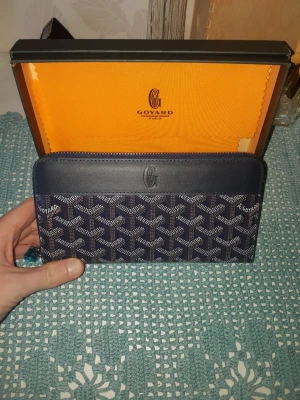 Mörkblå Goyard plånbok med mönster - Snygg mörkblå plånbok från Goyard med det klassiska vita och gråa Goyard-mönstret. Plånboken har dragkedja runtom och logga på framsidan. Tillverkad i canvas och läder, perfekt för dig som gillar exklusiva accessoarer.