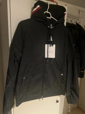 Moncler Windbreaker  - Snygg svart vindjacka från Moncler med dragkedja framtill, två fickor med tryckknappar och justerbar huva med vita och röda detaljer. Jackan har elastiska muddar vid ärmsluten och en Moncler-logga på ärmen. Perfekt för blåsiga dagar. Nfc scan. 10/10 skick!