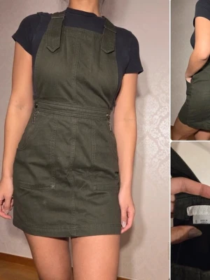 Olivgrön kort hängselklänning Urban Outfitters  - Snygg olivgrön hängselklänning med raka axelband och dragkedjor i midjan. Klänningen har två fickor fram och är gjord i ett kraftigare bomullstyg. Perfekt att styla med en t-shirt under för en cool och avslappnad look.