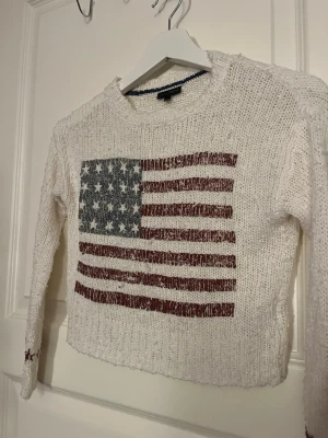 Vit stickad tröja med USA-flagga - Stickad vit tröja från Ralph Lauren med en stor amerikansk flagga på bröstet. Tröjan har lång ärm och ribbade muddar. 