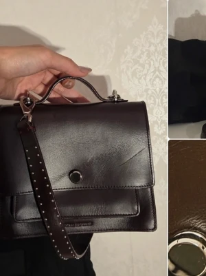 Mörkbrun handväska i läder från Royal Republiq - Snygg mörkbrun handväska i äkta läder med silverfärgade detaljer och ett dekorativt axelband med nitar. Väskan har ett stilrent, fyrkantigt format och ett lock med knappstängning. Perfekt för dig som gillar klassisk men edgy stil. Finns en repa vid framsidan, därav priset