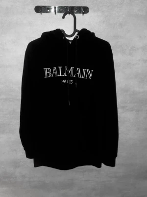Svart Balmain hoodie med tryck - Svart hoodie från Balmain med vit text 'BALMAIN PARIS' på bröstet. Klassisk modell med huva och dragsnören samt ribbade muddar vid ärmslut och nederkant. Perfekt för en stilren och trendig look.