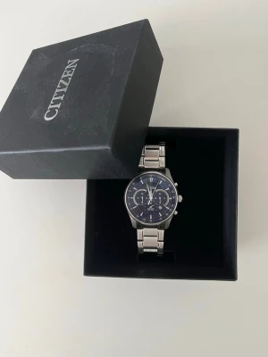 Citizen silverfärgad kronografklocka - Stilren kronografklocka från Citizen med silverfärgad länk i metall och mörkblå urtavla. Klockan har flera små urtavlor och tydliga markeringar, vilket ger en sportig men ändå klassisk look. Perfekt accessoar för dig som gillar detaljer och kvalitet.