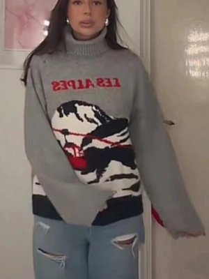 Grå stickad tröja med Les Alpes-tryck - Säljer en grå stickad tröja med hög krage och motiv av berg och linbana samt texten 'LES ALPES' i rött. Tröjan har långa ärmar och avslappnad passform. Perfekt för dig som gillar coola vintervibbar och statement-plagg.