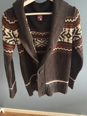 Brun kofta med mönster från the clockhoues  - Mysig brun kofta från the clockhoues med sjalkrage och knäppning framtill. Koftan har ett klassiskt nordiskt mönster i vitt, orange och beige över bröstet och ärmarna. Långärmad och ribbstickad vid ärmslut och nederkant. Perfekt för kalla dagar.
