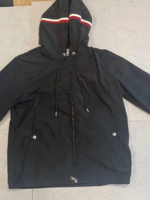 Svart Moncler vindjacka med huva - Svart Windbracker jacka från Moncler med huva och dragsko. Jackan har dragkedja framtill, två fickor med tryckknappar och Moncler-logga på ärmen. Huvan har en snygg röd, vit och blå rand. Perfekt för blåsiga dagar. Även kallad en wind breaker 