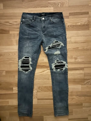 Amiri skinny jeans med hål och skinn  - Säljer ett par blå skinny jeans från Amiri med stora slitningar och hål över knäna. Jeansen har klassisk femficksdesign, dragkedjegylf och en cool, trendig look med mycket slitna detaljer. Perfekta för dig som gillar streetstyle och edgy mode.