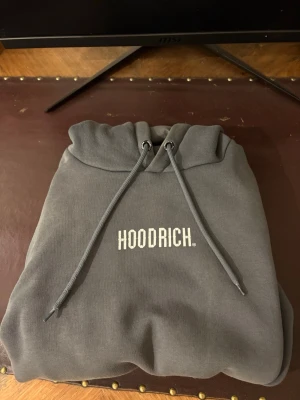 Grå hoodie från Hoodrich - Snygg grå hoodie från Hoodrich med vit broderad logga på bröstet. Klassisk modell med huva och dragsnören. Mjuk insida och ribbade muddar. Perfekt för en avslappnad streetwear-look.