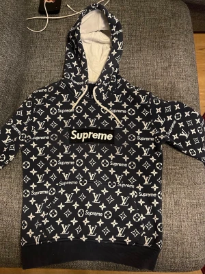 Supreme x  hoodie blå/vit - Snygg hoodie i blå med vitt all-over Supreme och l-v-mönster. Stor huva med vita snören och magficka framtill. Ribbstickade muddar vid ärmslut och nederkant. Perfekt för dig som gillar streetwear och vill sticka ut.