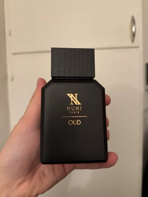 NUHI Paris OUD parfym - Elegant parfymflaska i matt svart med guldtext från NUHI Paris. Flaskan har en stilren, rektangulär form och ett stort svart lock med trästruktur. Doften är OUD, vilket ger en exklusiv och djup karaktär. 100ml av 100ml använd max 2 gånger nypris 2000.