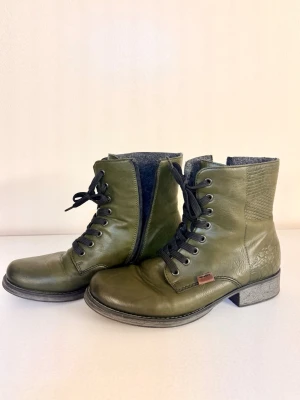 Rieker, olivgröna kängor med snörning - Snygga olivgröna kängor i läder med snörning och dragkedja på sidan. Kängorna har coolt mönstrad häl och skaft, samt robust sula i grått. Perfekta för dig som vill sticka ut med en unik stil.