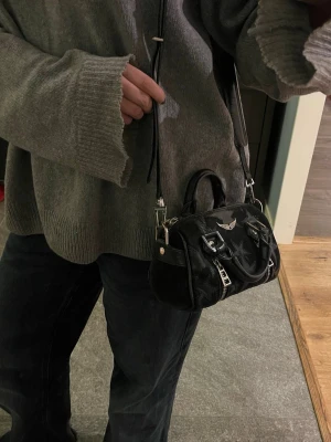 Svart handväska Zadig & Voltaire - Cool svart Sunny Nano handväska från Zadig & Voltaire med stjärnmönster, silverfärgade detaljer och dubbla dragkedjor framtill. Väskan har både handtag och justerbar axelrem med logotyp. Perfekt storlek för både vardag och fest. 