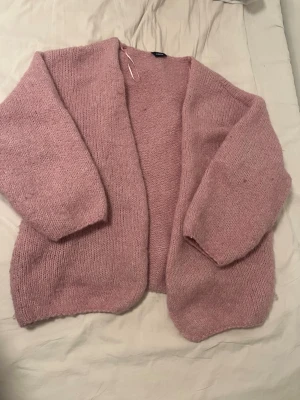 Rosa stickad kofta - Mysig rosa stickad kofta med öppen front och breda ärmar. Koftan har en avslappnad passform och är gjord i ett mjukt, fluffigt material som känns skönt mot huden. Perfekt att slänga på sig över en t-shirt eller topp för extra värme.