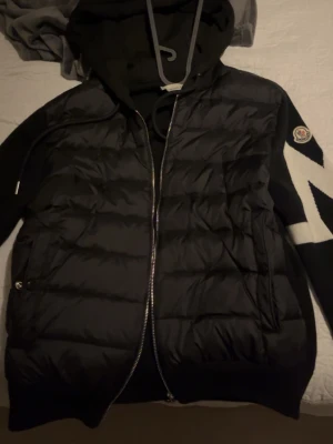 Svart dunjacka med luva - Säljer en svart dunjacka med stickade ärmar och stor vit M-detalj på ena ärmen. Jackan har dragkedja framtill, två fickor och en skön huva med dragsko. Perfekt för kyliga dagar och riktigt snygg streetstil.