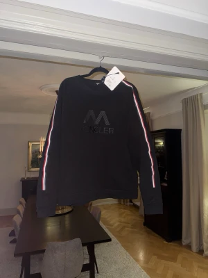 Svart Moncler sweatshirt med ränder - (KÖPT PÅ SICURO FASHION) Säljer en svart sweatshirt från Moncler med diskret logga på bröstet och vita/röda ränder längs ärmarna. Tröjan har rund halsringning och långa ärmar. Perfekt för dig som gillar stilrena och sportiga plagg.
